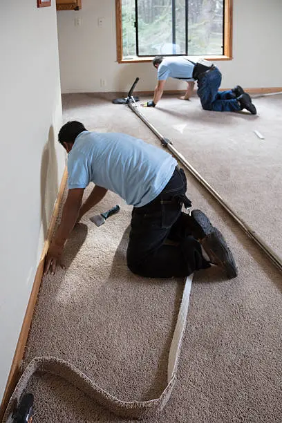 Two workers installing carpet in a room, kneeling and fitting edges.
