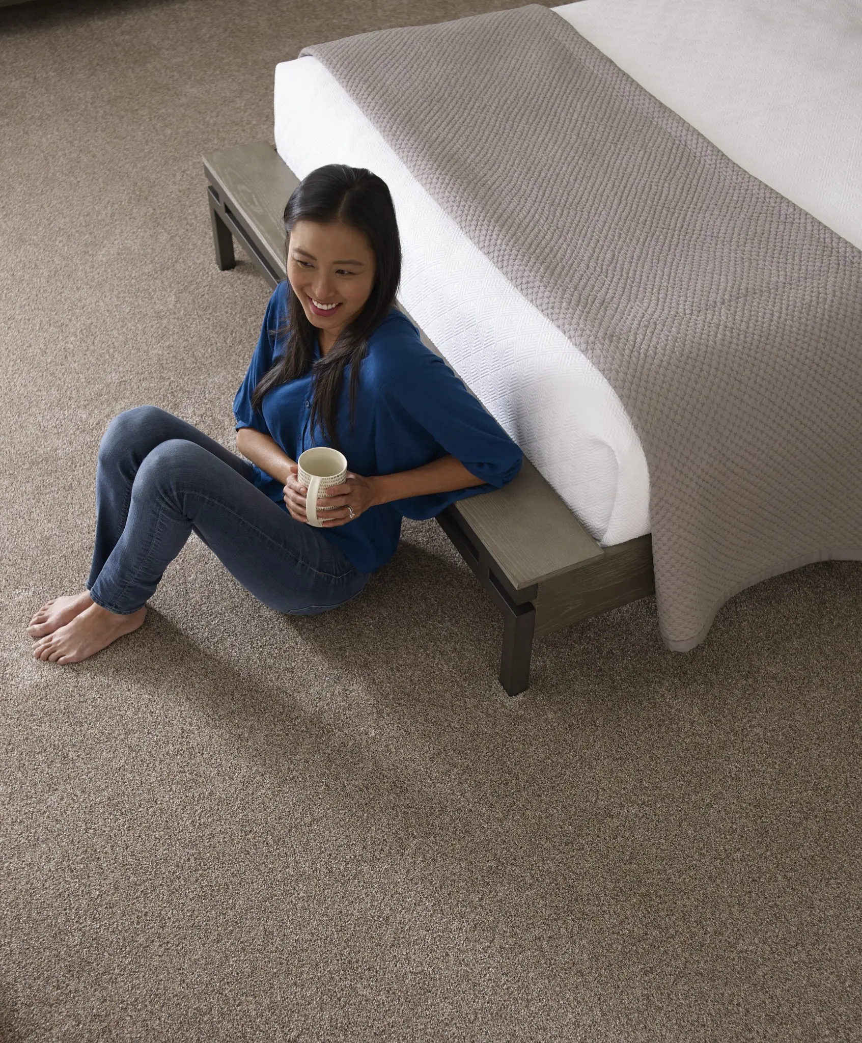 Woman sitting on carpeted floor, leaning against bed, holding cup.